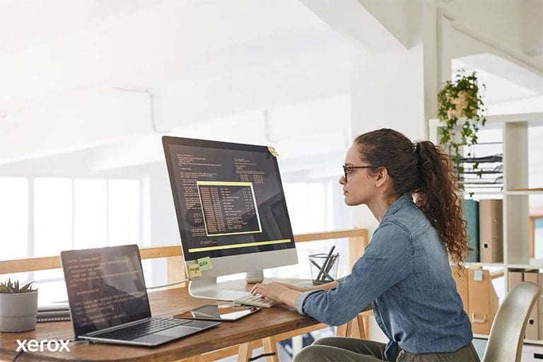 Une femme &agrave; son bureau, occup&eacute;e avec un ordinateur et un ordinateur portable, d&eacute;montrant sa productivit&eacute; dans un espace de travail moderne.