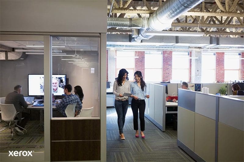 Deux femmes se prom&egrave;nent dans un espace de bureau ouvert moderne, engag&eacute;es dans une conversation et entour&eacute;es de postes de travail.