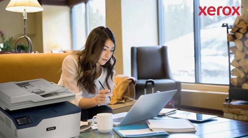 Une femme travaille sur un ordinateur portable dans un bureau ou un salon confortable, avec une imprimante Xerox compacte visible au premier plan, indiquant une int&eacute;gration transparente de l'impression avec les flux de travail num&eacute;riques.